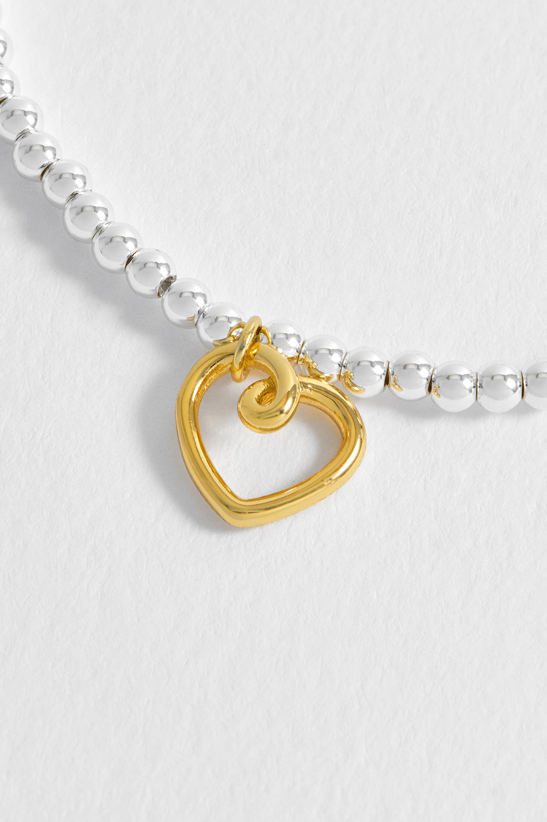 Gold heart-shaped charm on a silver beaded bracelet against a white background