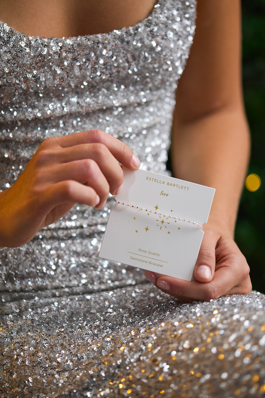 Person in a silver sequin dress holding a card with 'Estella Bartlett' branding.