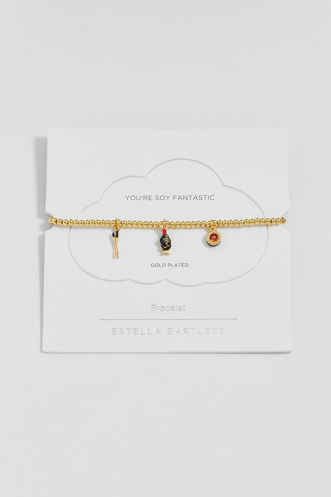A gold-plated beaded bracelet with three small charms (chopsticks, soy pack and a sushi), displayed on a white cloud-shaped card labeled 'YOU'RE SOY FANTASTIC' and 'ESTELLA BARTLETT'.