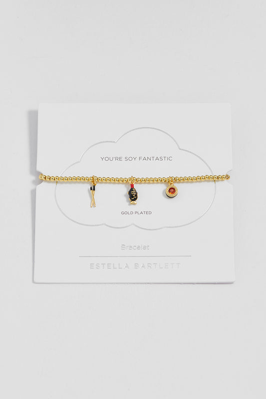 A gold-plated beaded bracelet with three small charms (chopsticks, soy pack and a sushi), displayed on a white cloud-shaped card labeled 'YOU'RE SOY FANTASTIC' and 'ESTELLA BARTLETT'.