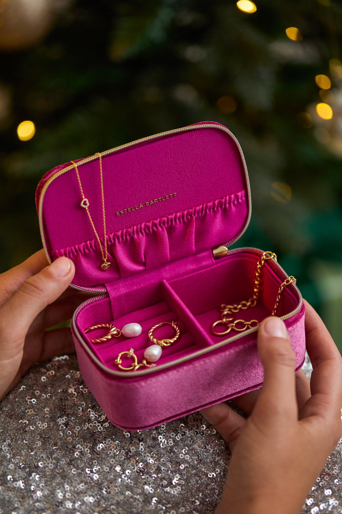 Pink jewelry case with gold jewelry held by hands against a blurred festive background